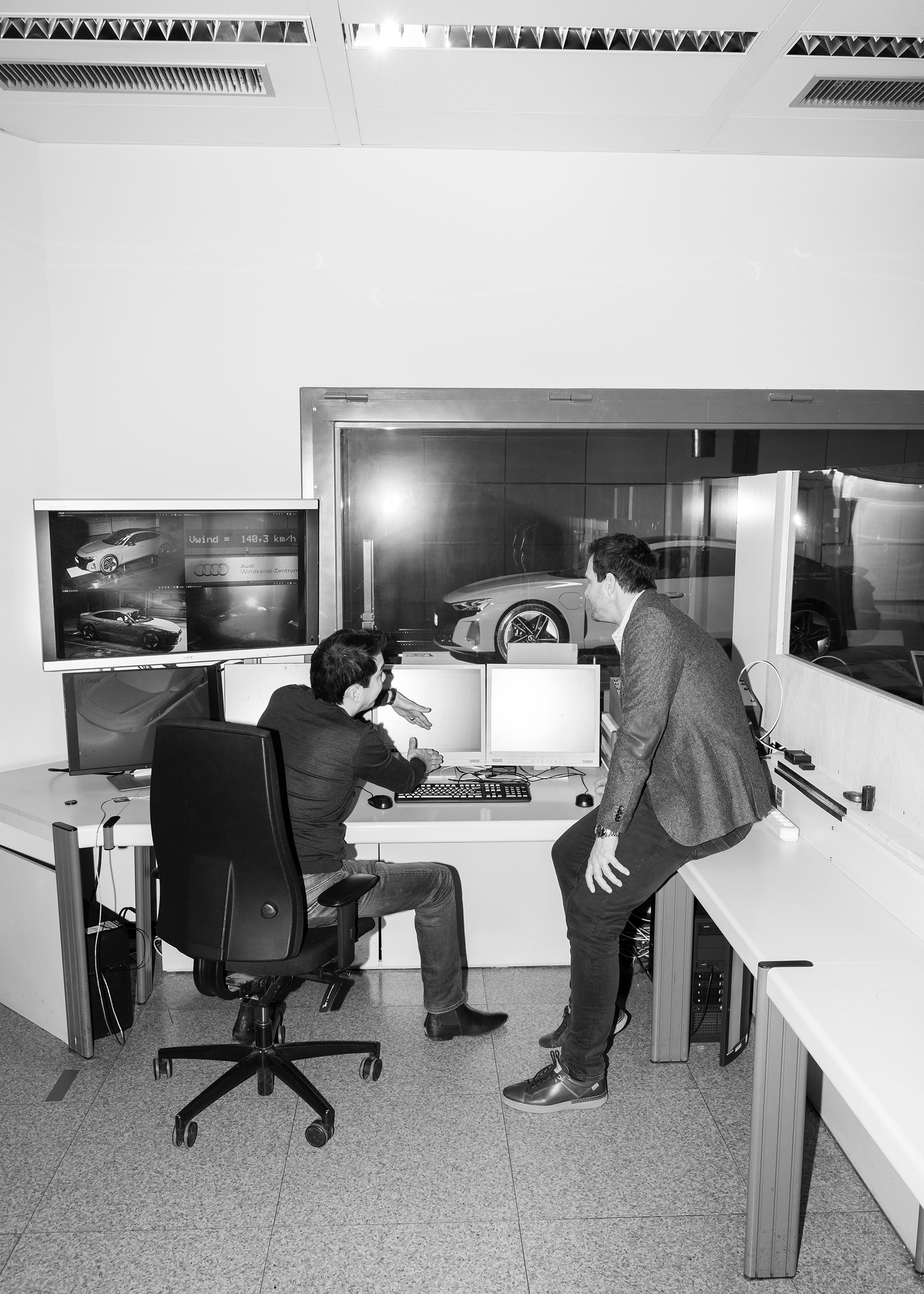 El Dr. Kentaro Zens y Thomas Redenbach conversan frente a varias pantallas de ordenador, con el Audi RS e-tron GT{ft_rs-e-tron-gt} detrás de una ventana.